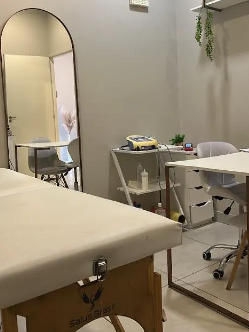Sala de atendimento com maca, espelho em arco, aparelho sobre o carrinho e prateleira com planta