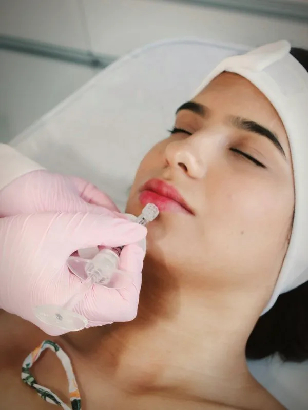 Profissional de luvas rosa aplicando preenchimento nos lábios de uma cliente deitada, de olhos fechados